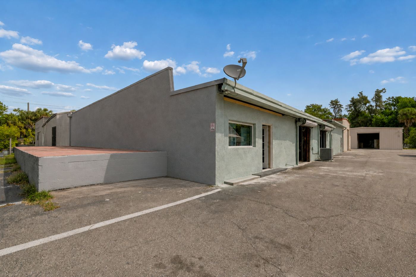 Current condition: gray weathered elevation, bare planter wall, cracked asphalt and aged outbuilding